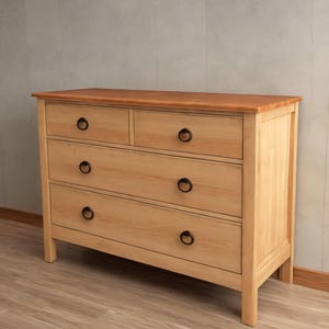 Op de afbeelding: Een lichte houten commode met vier lades en zwarte metalen handgrepen. De commode heeft een licht verweerde afwerking.