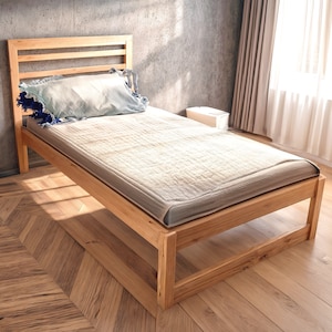 Könnte beinhalten: Ein Holzbettgestell mit hellbrauner Oberfläche. Das Bett hat ein Lattenkopfteil und ein schlichtes, rechteckiges Design. Das Bett ist aus massivem Holz gefertigt und hat eine natürliche, rustikale Optik.