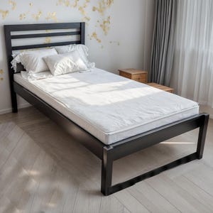 May include: A black wooden bed frame with a mattress and pillows. The bed has a headboard with horizontal slats. The bedding is white. The room has light-coloured wood flooring and a white wall with gold leaf accents.