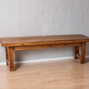 May include: A rustic wooden bench with a natural brown finish. The bench has a rectangular top and four legs. The bench is made of solid wood and has a simple, elegant design.