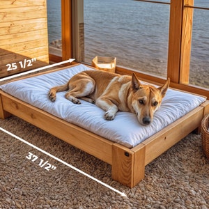 Pode incluir: Uma cama de cachorro de madeira com um colch&atilde;o branco, medindo 95,25 cm por 64,77 cm e 15,24 cm de altura. Um cachorro marrom est&aacute; deitado na cama.