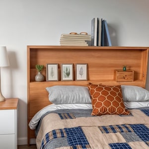May include: A wooden headboard with a built-in shelf and three framed botanical prints. The bed is made with a white sheet, two gray pillows, and a brown patterned pillow. A blue and beige patchwork quilt covers the bed.