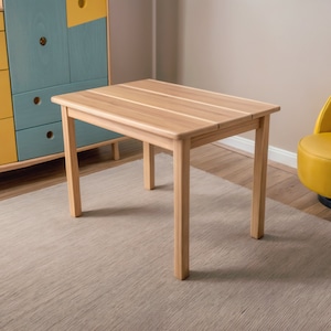 Puede incluir: Una mesa de madera clara con una tapa rectangular y cuatro patas. La mesa está sobre una alfombra beige en una habitación con una silla amarilla y un armario colorido.