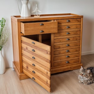 May include: A wooden cabinet with multiple drawers, some of which are open. The cabinet is made of light brown wood and has a rustic look. The drawers have metal handles. A cat is sitting on the floor in front of the cabinet.