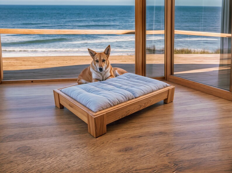 Pode incluir: Uma cama para c&atilde;es azul claro com uma moldura de madeira est&aacute; em um piso de madeira. Um c&atilde;o marrom e branco est&aacute; sentado na cama, olhando para a c&acirc;mera. A cama est&aacute; na frente de uma janela grande com vista para o oceano.