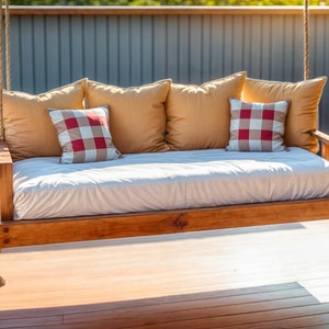 May include: A wooden swing bed with a white mattress and yellow and red patterned pillows. The swing is hanging from a wooden frame and is suspended over a wooden deck.