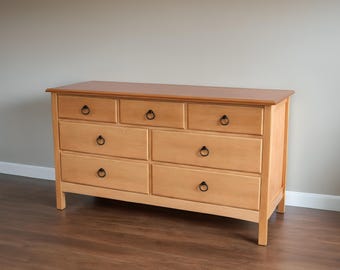 May include: A light wood dresser with six drawers and black ring pulls. The dresser has a slightly rounded top and sits on a wood floor.