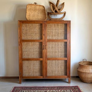 Puede incluir: Un armario de madera con dos puertas y paneles de ratán tejido. El armario está sobre una alfombra con un patrón floral. Hay dos cestas tejidas en la parte superior del armario.