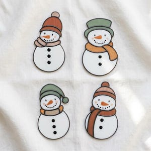 May include: Four stained glass snowman ornaments featuring white bodies with black button details, orange carrot noses, and dotted smiles. Each snowman wears a unique hat and scarf combination in shades of muted orange, green, and brown.