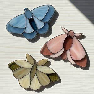 May include: Three stained glass moth decorations in shades of blue, pink, and cream with metallic borders. The moths have layered wings and detailed body shapes, ideal for home decor or as gifts.