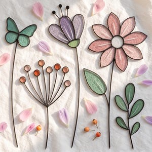 May include: A collection of stained glass garden stakes featuring a teal butterfly, a purple flower, a pink daisy with a green leaf, a branch with green leaves, and a dandelion-like plant with orange spheres, all accented by scattered petals.