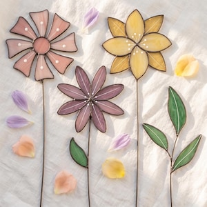 May include: Three stained glass flowers on thin metal stems, including a light pink flower, a dark purple flower, and a yellow flower, accompanied by a separate green stained glass leaf stem, all scattered with loose, colorful flower petals.