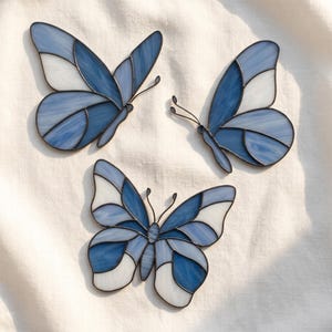 Puede incluir: Tres adornos de mariposa hechos de vidrieras, con paneles de vidrio blanco translúcido y varios tonos de azul. Cada mariposa está construida con líneas de soldadura de metal oscuro que delinean las alas e incluye antenas de alambre fino.