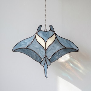 May include: A stained glass suncatcher shaped like a manta ray, featuring marbled light blue glass panels and two opaque white glass sections near the top. The pieces are joined by dark metal solder lines and it hangs from a thin metal chain.