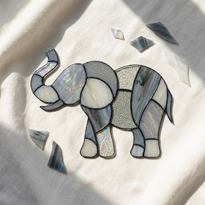 Puede incluir: Una figura de elefante de vitral construida con varias tonalidades de vidrio gris, blanco e iridiscente unidas por líneas de soldadura oscuras. El elefante se muestra de perfil con la trompa levantada, rodeado de varios fragmentos de vidrio.
