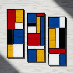 May include: Three rectangular stained glass panels with a Mondrian-inspired design. Each panel features a grid of geometric shapes in red, yellow, blue, black, and white, framed in black. The panels are arranged on a light wood surface.