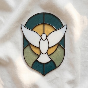 May include: An oval stained glass panel featuring a white dove with outstretched wings centered against a yellow circular background, surrounded by segments of dark teal and muted green glass, all held together by dark metal lead lines.