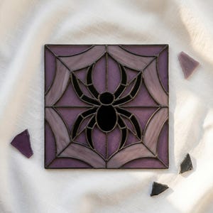 May include: A square stained glass panel featuring a black spider centred within a web design. The glass pieces are in varying shades of purple and light lavender, held together by dark metal solder lines. Small glass shards lie nearby.