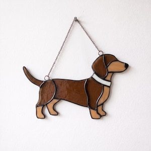 Op de afbeelding: Een glas-in-lood suncatcher in de vorm van een teckel. Het object bestaat uit donkerbruine en lichtbeige glazen segmenten verbonden door donkere metalen soldeerlijnen. Een witte glazen halsband is zichtbaar rond de nek met een metalen ketting.