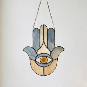 May include: Stained glass Hamsa hand with a central eye design. The hand features a color palette of blue, cream, and amber, with a brass-colored metal frame and chain for hanging. The Hamsa is a symbol of protection and good fortune.