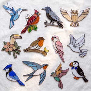 May include: A collection of twelve stained glass bird ornaments featuring a blue hummingbird, a red cardinal, a black crow, a tan owl, a toucan, a robin, a pink parrot, a white dove, a blue jay, a swallow, a small colorful bird, and a puffin.