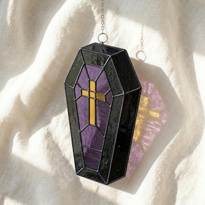Puede incluir: Un atrapasoles de vitral con forma de ataúd que presenta bordes de vidrio negro opaco, un centro de vidrio morado translúcido y una cruz central de vidrio de color dorado. La pieza cuelga de una fina cadena metálica sujeta a una argolla.