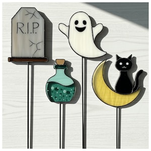 Può includere: Set di picchetti da giardino a tema Halloween. Include una lapide con "R.I.P.", un fantasma sorridente, una bottiglia di pozione e un gatto nero su una luna crescente. Realizzati in vetro colorato con steli in metallo nero.