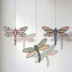 May include: Three stained glass dragonfly ornaments hanging from silver chains. The left dragonfly has pink and pale green wings with a pink body. The center one features green and beige wings with a green body. The right one has pink and green wings with a blue body.