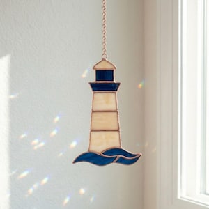 May include: Stained glass lighthouse with a cream-colored body, navy blue accents, and a blue wave base. The lighthouse is suspended by a copper chain. A decorative nautical-themed item.