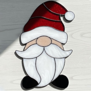 Puede incluir: Gnomo de vidriera con gorro rojo, barba blanca y zapatos negros. El gorro del gnomo tiene un ribete blanco y una bola blanca en el extremo. La cara del gnomo es de color marrón claro.