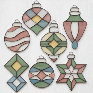 May include: A collection of seven stained glass Christmas ornaments in various shapes. The ornaments feature pastel colors like pink, green, and blue, with gold glitter accents. Each ornament has a metal loop for hanging.