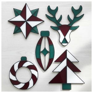 Op de afbeelding: Een verzameling glas-in-lood kerstversieringen in diverse vormen: een ster, een rendierkop, een ornament, een krans en een kerstboom. De ornamenten hebben een kleurenschema van wit, bordeauxrood en groen met zwarte contouren.