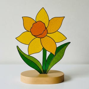 May include: A stained glass daffodil sculpture with yellow petals and an orange center. The flower has green leaves and a green stem, set on a round wooden base. The daffodil is a spring-themed decorative item.