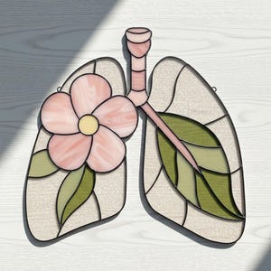 Könnte beinhalten: Buntglas-Lungen-Design mit einer rosa Blume und grünen Blättern. Das Lungendesign besteht aus verschiedenen Glasteilen mit einem hellbeigen Hintergrund. Die Blume hat eine gelbe Mitte. Das Stück ist schwarz umrandet.