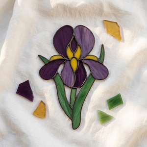 May include: A stained glass iris flower suncatcher featuring deep purple, light purple, and bright yellow petals with green glass leaves, outlined in dark metal solder, accompanied by four loose, small, irregular glass shards in purple, yellow, and green.