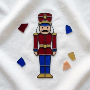 May include: A stained glass nutcracker figure featuring a red jacket and hat, blue trousers, and yellow accents, outlined in dark metal solder. The figure is surrounded by five small, loose, multi-coloured glass shards in red, blue, yellow, and peach.