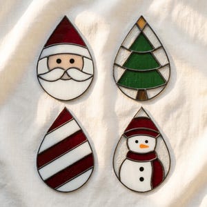 May include: Four teardrop-shaped stained glass ornaments featuring holiday designs. One depicts a Santa Claus face, one a green Christmas tree, one a red and white candy cane striped pattern, and one a snowman wearing a red hat and scarf.