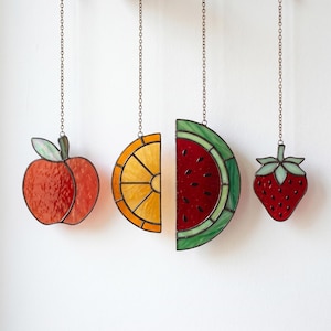 Op de afbeelding: Vier hangende fruitornamenten van glas-in-lood aan metalen kettingen. De set bevat een ronde oranje appel met groene blaadjes, een gele sinaasappelschijf, een rood watermeloenpartje met groene schil en een rode aardbei met groene blaadjes.