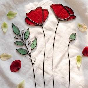 Peut inclure: Trois tiges décoratives en vitrail comprenant deux fleurs de coquelicot rouge vif avec des soudures en métal sombre, une branche à feuilles vertes avec des accents blancs et un bouton vert sur une fine tige métallique, posées sur un tissu blanc.
