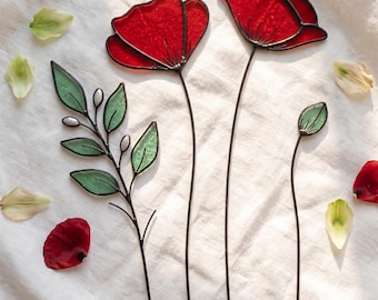 May include: Three stained glass decorative stems featuring two vibrant red poppy flowers with dark metal soldering, one green leafy branch with small white accents, and a single green bud on a thin metal wire stem, arranged on a white fabric.