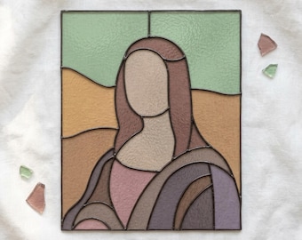 Puede incluir: Un panel rectangular de vidriera que presenta un retrato estilizado y sin rostro inspirado en la Mona Lisa. La composición utiliza piezas de vidrio texturizado en tonos verde salvia, beige, marrón, malva y morado, unidas por líneas de soldadura oscuras.