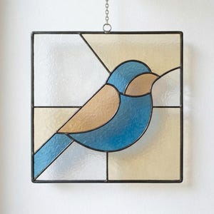 May include: Stained glass square panel featuring a blue and tan bird design. The bird is set against a background of white and pale yellow glass, framed by a black metal border. A chain is attached for hanging.