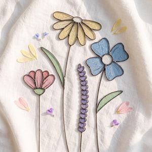 May include: Four stained glass flower stems featuring a yellow daisy, a pink blossom, a purple lavender-like spike, and a blue flower. Each piece is crafted with dark metal soldering and includes scattered loose glass petals in matching colors.