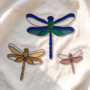 May include: Three dragonfly ornaments crafted from stained glass with dark metal soldering. The largest features blue and green wings, the medium one is amber-coloured, and the smallest is light pink. Each has a segmented body and four wings.