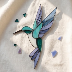 May include: A stained glass hummingbird suncatcher featuring a palette of teal, light blue, lavender, and white glass segments joined by dark metal solder lines, accompanied by several small loose triangular glass pieces in matching colours.
