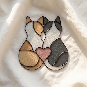 Op de afbeelding: Een glas-in-lood suncatcher met twee katten die naast elkaar zitten, verbonden door een centraal roze hart. De ene kat is van getextureerd amberkleurig en wit glas, de andere van antracietgrijs en wit glas, omlijnd met donker metaalsoldeer.