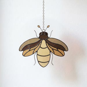 May include: A stained glass bee suncatcher featuring a segmented body and wings in shades of cream, tan, and dark brown. The insect is constructed with dark metal solder lines and is suspended from a silver-toned metal chain for hanging display.