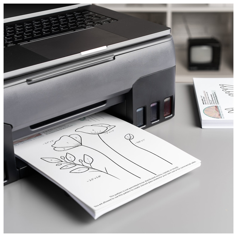 May include: A black and silver inkjet printer with a white sheet of paper with a flower design and numbers printed on it. The paper is being fed into the printer. The text on the paper reads "This pattern is not permitted to be distributed, copied or resold. You are allowed to use this pattern to create finished works for your own use or for commercial sale."