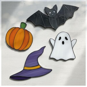 May include: A selection of Halloween-themed stained glass decorations. Features a black bat, an orange pumpkin, a white ghost, and a purple witch's hat with a yellow band. Each piece is outlined in black.