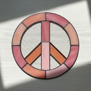 May include: A stained glass peace sign in shades of pink, peach, and orange. The symbol is round with a clear, textured center and a black border. The peace sign is a classic symbol of peace and harmony.